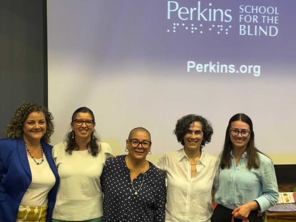 Cinco mujeres sonríen y posan para una foto en frente de una pantalla de presentación. La pantalla es de color azul y tiene el logo de Perkins, y también su sitio web de Perkins.org.