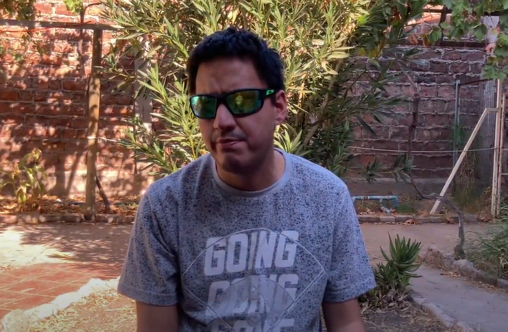 Un chico en frente de un arbol con lentes oscuros y una camiseta gris.