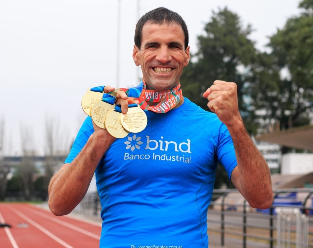 Un hombre en una camiseta azul sonríe con cuatro medallas de oro en su cuello. Su camiseta dice "Bind Banco Industrial"