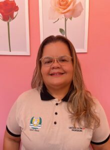 Uma mulher de cabelo longo e óculos, sorrindo, está vestindo uma camisa polo bege com o texto bordado "LUCIOLA ALVINO PROFESSORA." Ela está de pé em frente a uma parede rosa com quadros.