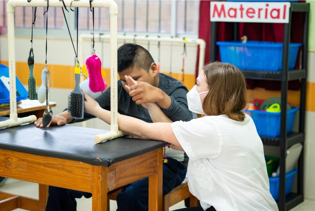 Una docente con una mascarilla está ayudando a un estudiante en usar objetos sensoriales colgando de un marco en una clase.