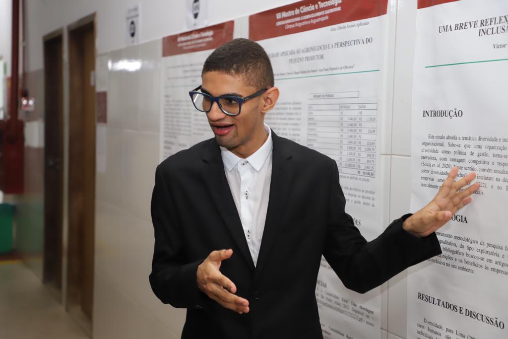 Uma pessoa com cabelo curto e óculos, vestindo um terno preto e camisa branca, fala animadamente enquanto gesticula com as duas mãos. Ela está de pé em frente a um quadro de avisos cheio de gráficos e textos impressos.