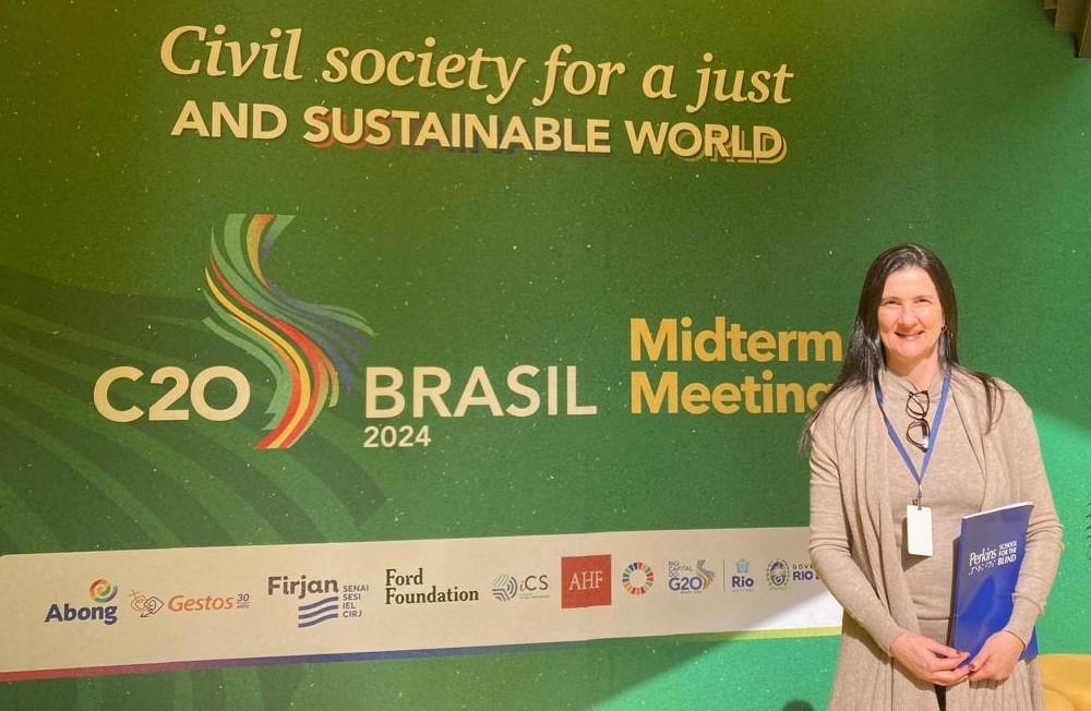 Una mujer sonríe frente a un fondo verde con el texto "La sociedad civil por un mundo justo y sostenible". El telón de fondo también dice "Reunión de mitad de período C20 Brasil 2024" y muestra varios logotipos de organizaciones. Ella sostiene una carpeta azul y porta una insignia de la conferencia.