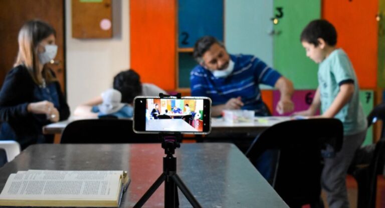 Un smartphone sobre un trípode graba una escena de clase en la que un profesor con mascarilla ayuda a los niños con sus actividades en una mesa. Hay taquillas de colores al fondo y un libro abierto en un pupitre en primer plano.