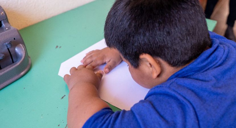 Un niño con una camiseta azul se inclina sobre una mesa verde, concentrado en doblar o interactuar con un pedazo de papel blanco. Un dispositivo gris es parcialmente visible en el borde de la mesa.