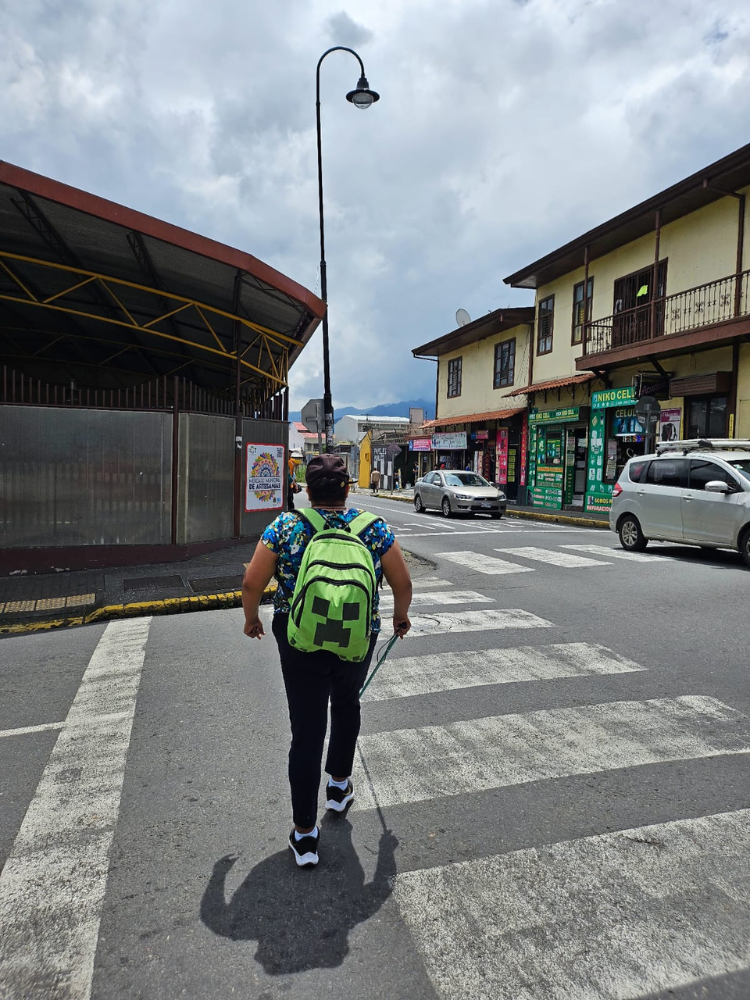 Foto de una mujer con una mochila verde usando un bastón verde para cruzar la calle.