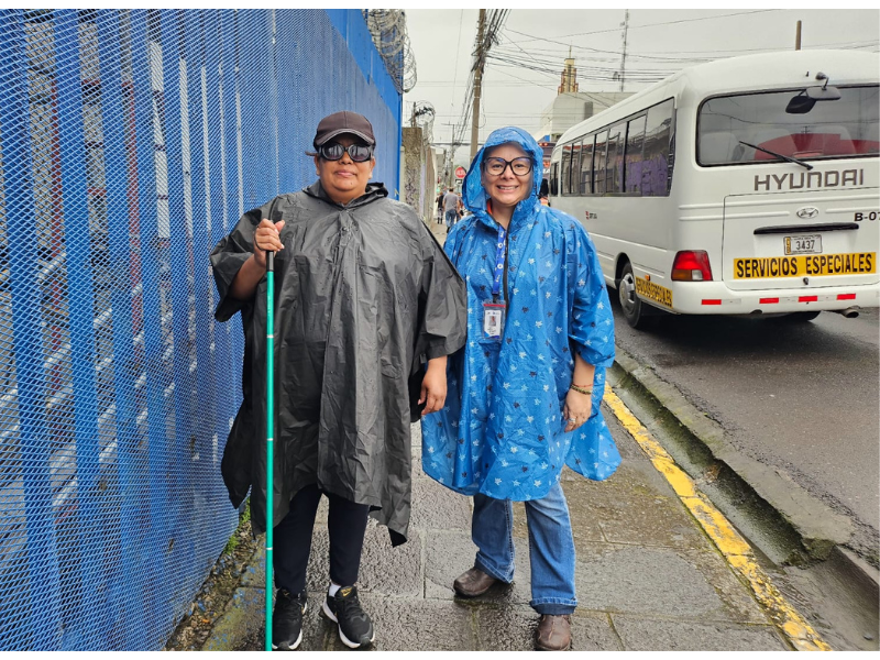 Foto de dos mujeres en la calle, la mujer de la izquierda trae un impermeable negro con un bastón verde y la mujer del lado derecho trae un impermeable azúl con estrellas y está sonriendo.