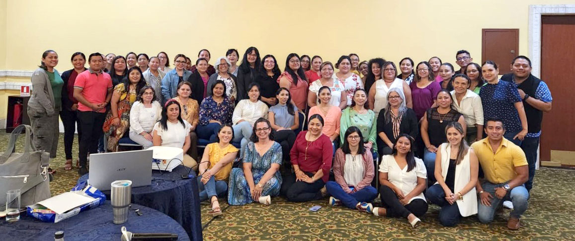 Un gran grupo de personas reunidas en el interior para una foto de grupo. Están sonrientes y vestidos de manera informal, de pie y sentados en varias filas. En primer plano se ven un proyector y algunas mesas.