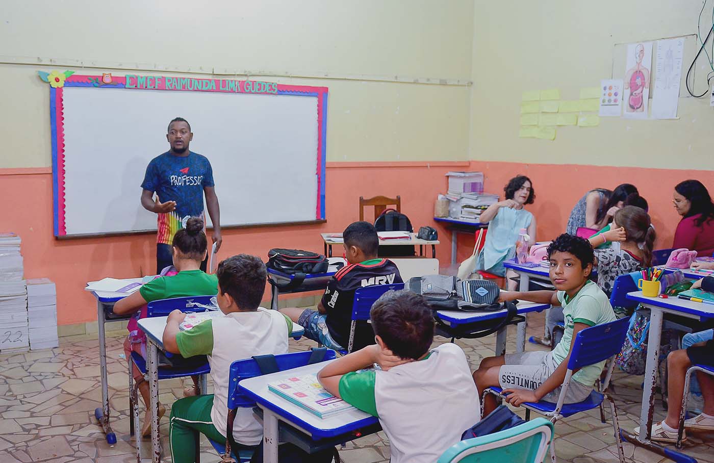 Un profesor está de pie delante de un aula con alumnos sentados en pupitres. Las paredes son de colores y los alumnos llevan uniforme