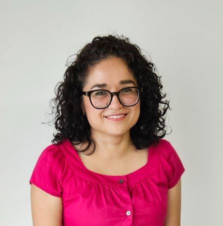 Foto de una mujer sonriendo en un fondo blanco, con lentes y una blusa rosa.