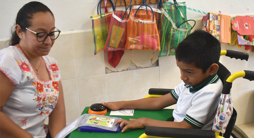 Un niño en silla de ruedas, con uniforme escolar, señala un libro sobre una mesa. Una mujer le observa sentada cerca. En la pared del fondo cuelgan bolsas y materiales de colores.