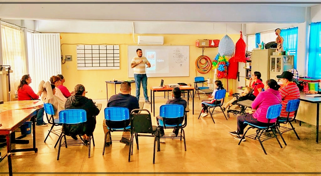 Saúl se sitúa frente a un grupo de adultos sentados en un aula. Un proyector muestra contenidos en una pantalla detrás de él. La sala es luminosa, con una decoración colorida y materiales educativos. Los asistentes escuchan atentamente.