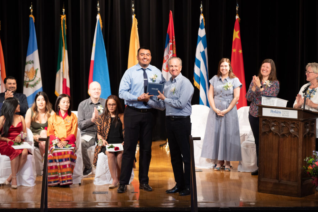 Saul, un hombre alto, recibe un premio en el escenario de manos de otro hombre ante el público. Ambos sonríen mientras posan para una foto. Detrás de ellos, varias personas están sentadas, con banderas de varios países desplegadas en el fondo