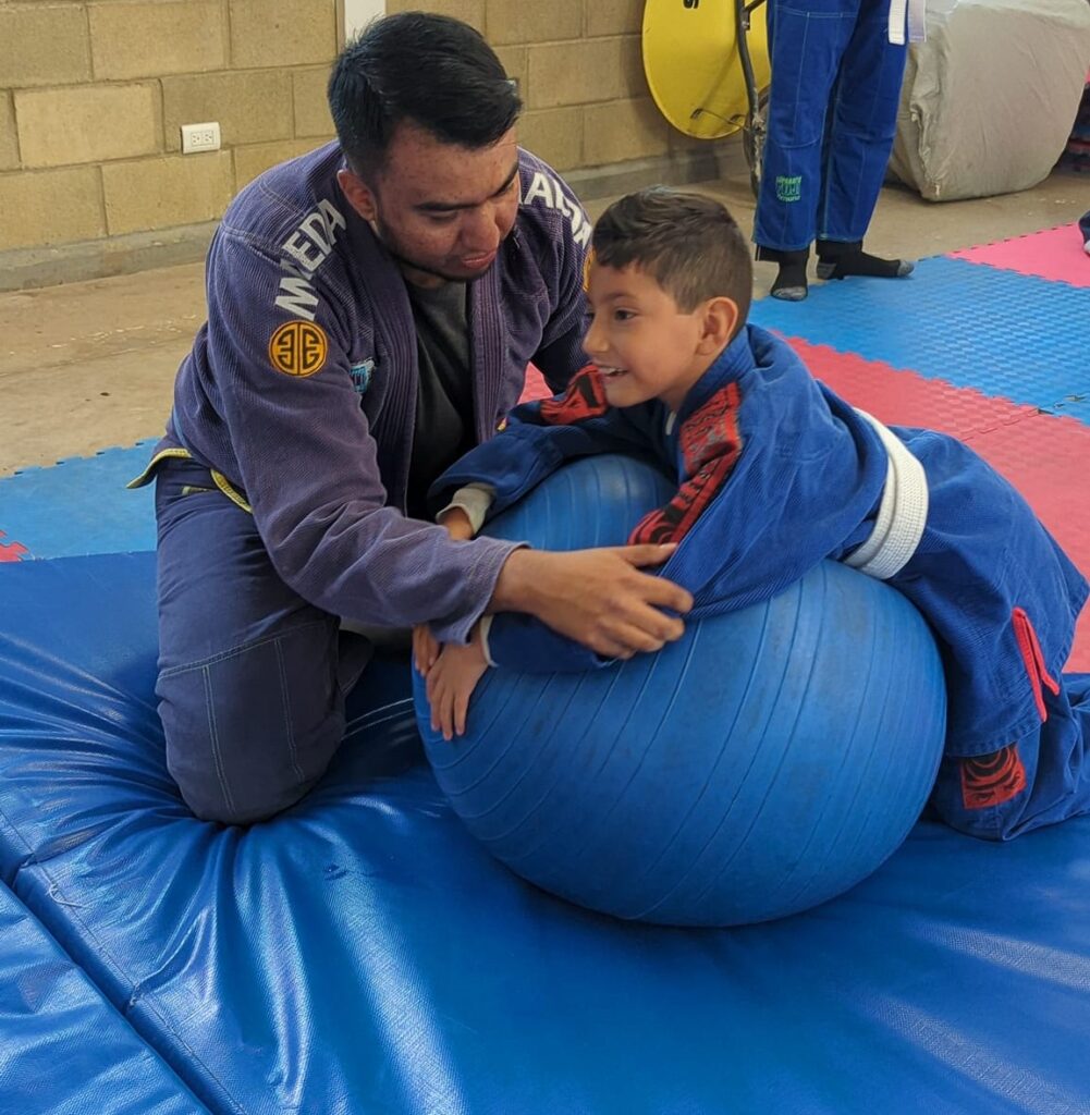 Un hombre y un niño con uniforme de artes marciales practican con una pelota azul. El chico, sonriente, se inclina hacia delante sobre la pelota mientras el hombre le sostiene. Están sobre una colchoneta azul en un gimnasio.