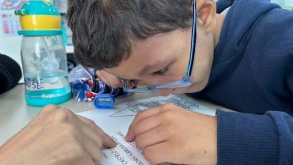 Un niño con cabello corto y oscuro, que lleva gafas, está leyendo atentamente un papel escrito a mano sobre una mesa. Se está inclinando hacia adelante, concentrándose intensamente. Hay una botella de agua y un estuche de lápices con crayones cerca, y se ve la mano de un adulto señalando el papel.