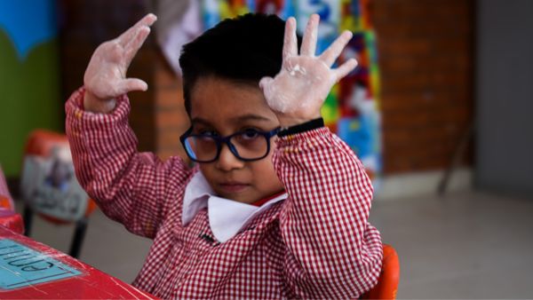 Un niño con gafas y camisa roja a cuadros levanta ambas manos, mostrando las palmas cubiertas de polvo blanco. El fondo tiene elementos coloridos y una pared de ladrillos difuminada. El niño mira directamente a la cámara.