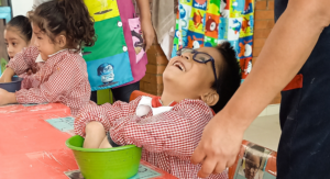 Un niño con camisa roja a cuadros y gafas se ríe mientras juega con un cuenco verde. En el fondo se ven otros niños y adornos de colores.