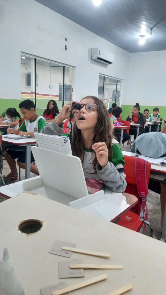 Un estudiante con el pelo largo mira a través de una pequeña lupa en un aula. Otros estudiantes con uniformes blancos y verdes están sentados en sus pupitres, concentrados en su trabajo. El aula tiene paredes de color verde claro.