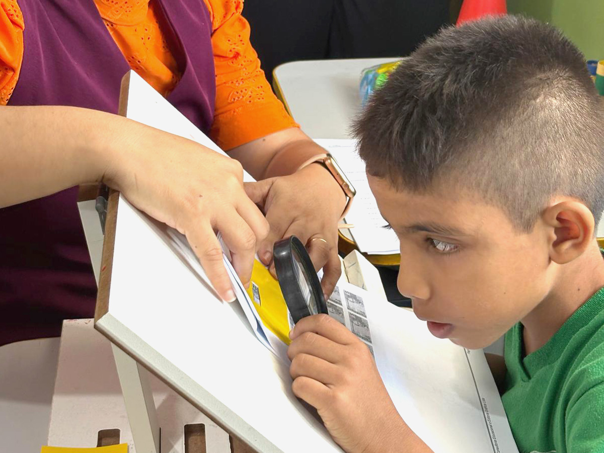 Un niño con una camiseta verde usa una lupa para mirar un trozo de papel sobre una superficie inclinada. Un adulto con una camiseta naranja y un chaleco violeta le ayuda, señalando el papel.