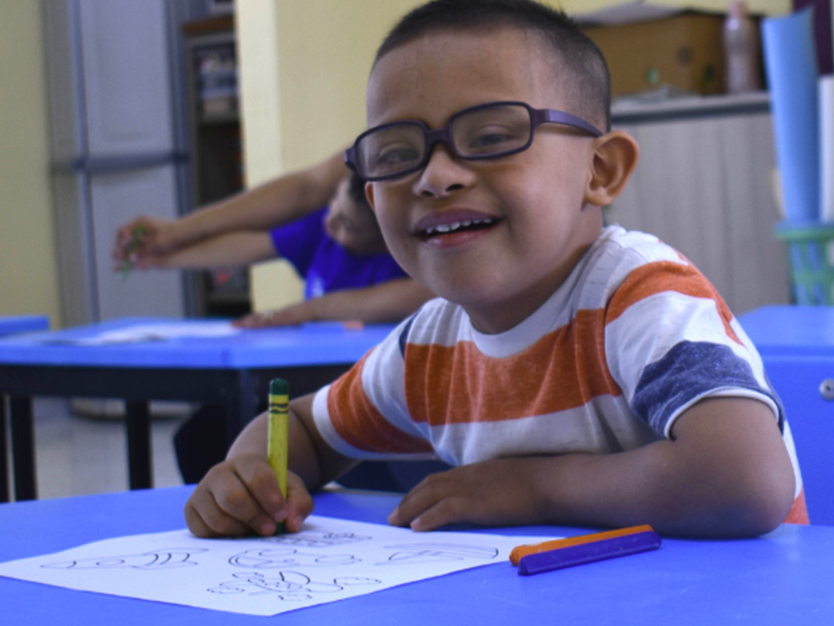 Un niño sonriente con gafas y una camiseta a rayas está sentado en una mesa azul y colorea con un crayón verde. El fondo muestra un aula con otro estudiante borroso en la distancia