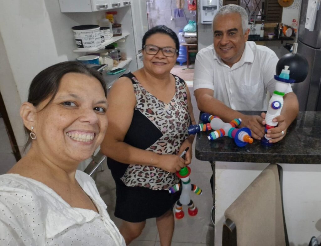 Três pessoas sorriem enquanto seguram brinquedos coloridos e cilíndricos em uma cozinha. Uma pessoa está tirando uma selfie. O balcão tem vários outros brinquedos. O fundo mostra prateleiras e eletrodomésticos da cozinha.