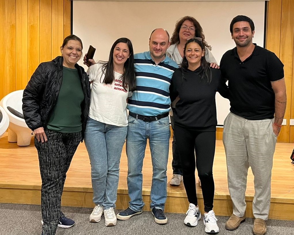 Un grupo de seis personas permanecen juntas, sonrientes, frente a una pared de madera y una pantalla. Visten de manera informal, algunos con chaqueta y zapatillas de deporte. Parecen estar en un ambiente relajado y amistoso.