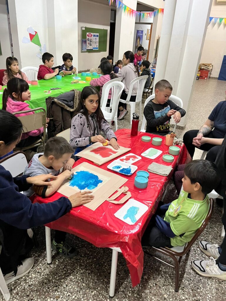 Niños y niñas sentadas alrededor de mesas cubiertas con paños rojos y verdes, realizando actividades de manualidades. Están utilizando papel y pintura, con algunos niños concentrados en sus creaciones. La sala está decorada con banderolas de colores.