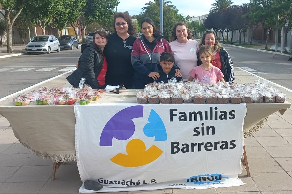 Un grupo de personas sonrientes, entre ellas dos niños, se para detrás de una mesa con una pancarta en la que se lee «Familias sin Barreras». La mesa está llena de productos empaquetados. Están al aire libre en una calle bordeada de árboles.