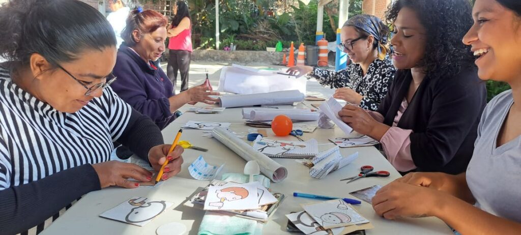 A group of people sitting around a table outdoors, engaging in arts and crafts. They are smiling and working on various projects with paper and other materials. Trees and traffic cones are visible in the background.