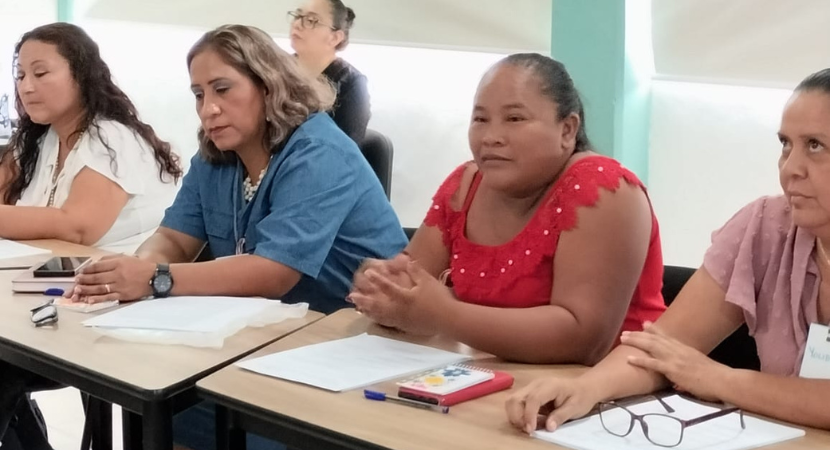 Un grupo de mujeres se sienta a la mesa durante una reunión o un taller. Tienen papeles y cuadernos delante y parecen atentas. El entorno es luminoso, con paredes claras y ventanas al fondo.