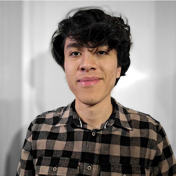 Persona sonriente con el pelo oscuro que lleva una camisa a cuadros negra y marrón, de pie sobre un fondo gris borroso.