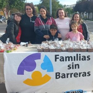 Un grupo de personas sonrientes, entre ellas dos niños, se para detrás de una mesa con una pancarta en la que se lee «Familias sin Barreras». La mesa está llena de productos empaquetados. Están al aire libre en una calle bordeada de árboles.