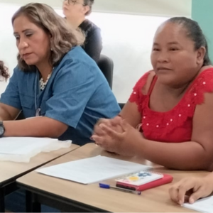 Un grupo de mujeres se sienta a la mesa durante una reunión o un taller. Tienen papeles y cuadernos delante y parecen atentas. El entorno es luminoso, con paredes claras y ventanas al fondo.