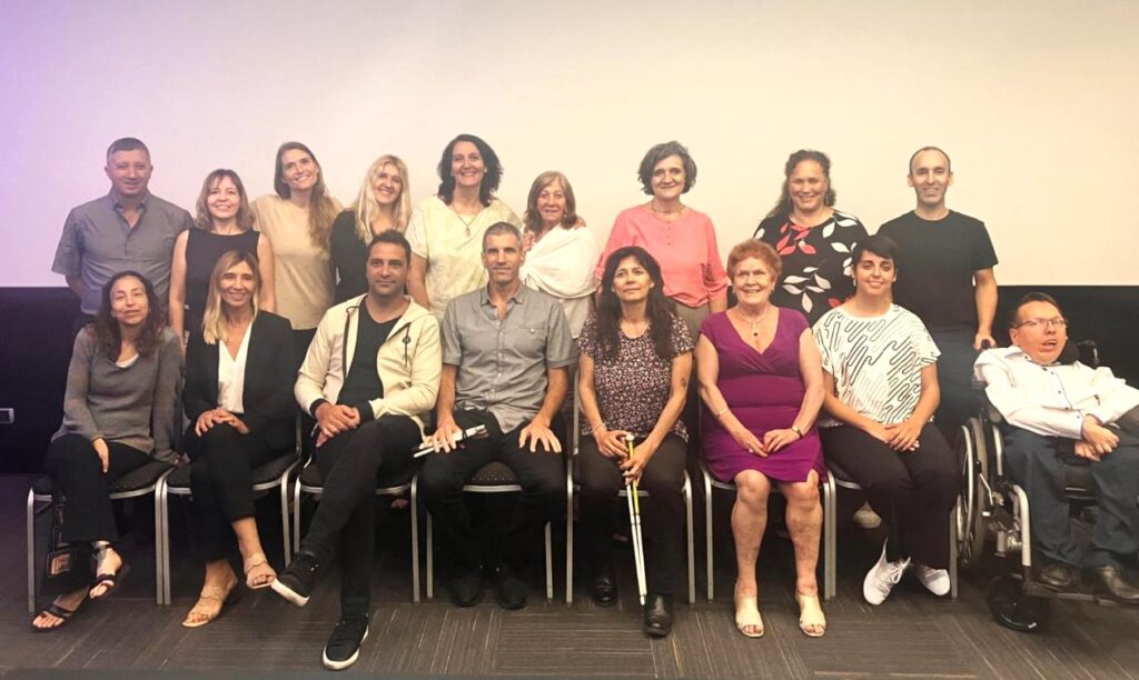 Un grupo de dieciséis adultos, incluidos hombres y mujeres, algunos con discapacidad, posan juntos en un espacio interior frente a una pared blanca, sonriendo a la cámara.