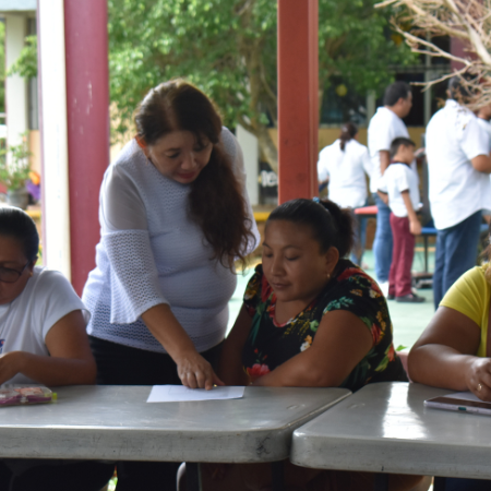 Una mujer ayuda a otras tres personas sentadas en una mesa con papeles, bajo un espacio cubierto al aire libre. Sobre la mesa hay un proyector. Al fondo, más gente se reúne cerca de unos árboles.