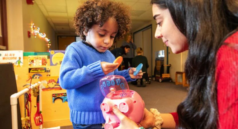 Valentina ELP Chile at Perkins Un niño pequeño con un suéter azul juega con monedas de juguete junto a una alcancía rosa, mientras un adulto con un suéter rojo le ayuda. Al fondo se ven libros y juguetes infantiles de colores.