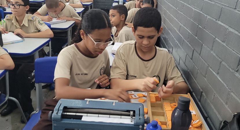 Dos estudiantes con uniforme beige se sientan en los pupitres de un aula, utilizando una máquina de escribir Perkins Braille y herramientas de aprendizaje táctil, mientras sus compañeros trabajan en pupitres cercanos. Sobre los pupitres se ven botellas de agua y bolsas.