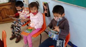 Tres niños pequeños están sentados en sillas naranjas en un aula, cada uno leyendo un libro colorido. Al fondo se ven una pizarra verde y un armario de madera.