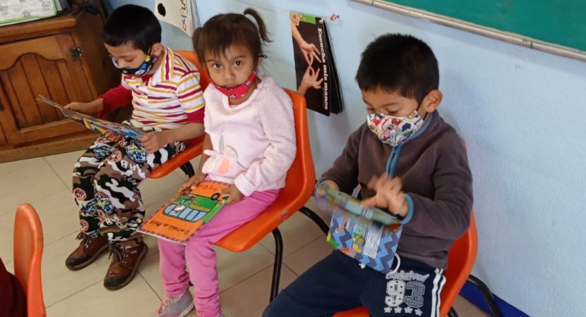Tres niños pequeños están sentados en sillas naranjas en un aula, cada uno leyendo un libro colorido. Al fondo se ven una pizarra verde y un armario de madera.