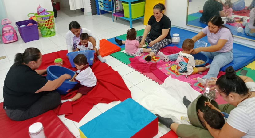 Varios adultos y niños pequeños se sientan sobre tapetes coloridos en una sala de juegos, participando en actividades de juego sensorial con diversos recipientes y materiales. Al fondo, se ven estantes con juguetes y materiales.