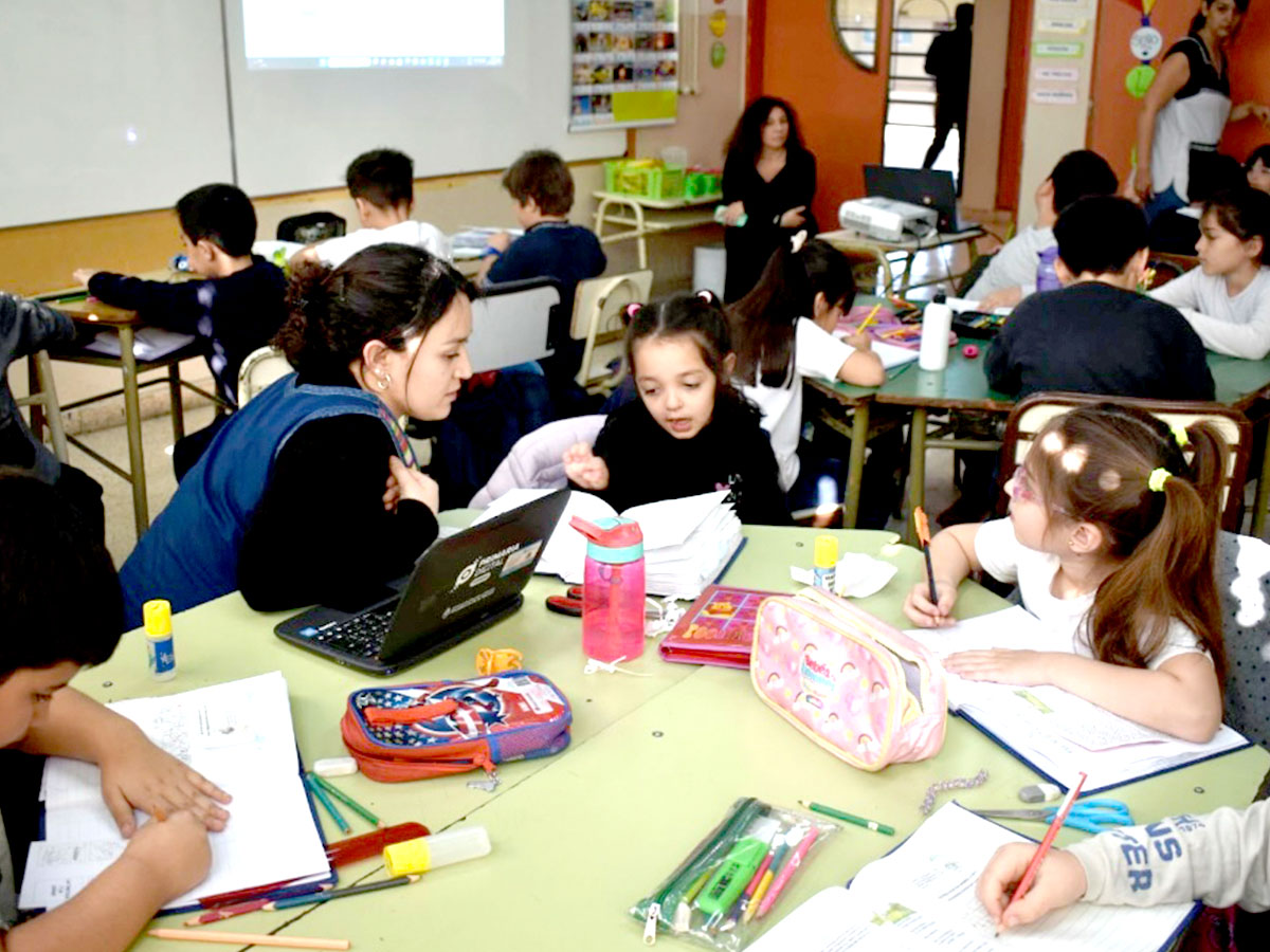 Un aula con estudiantes jóvenes sentados en mesas trabajando en sus tareas. Una profesora interactúa con una niña mientras otros estudiantes escriben, dibujan y usan útiles escolares. Se ven libros, estuches y botellas de agua sobre las mesas.