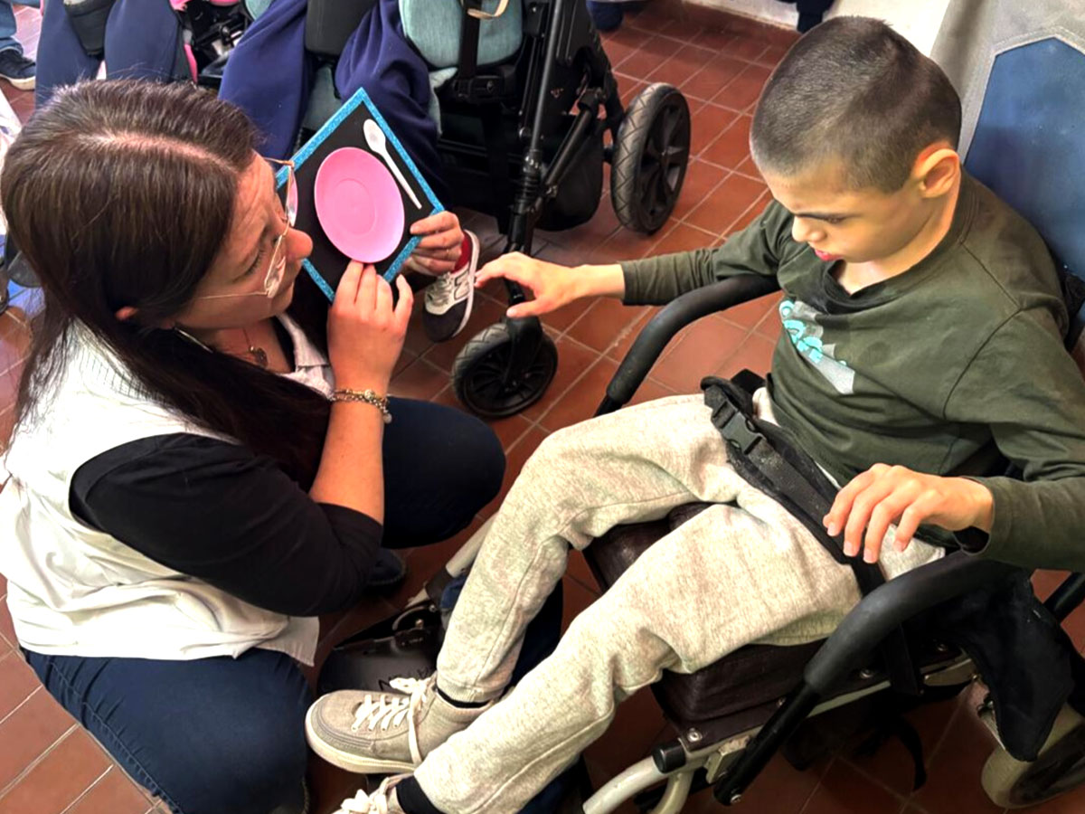 Una maestra se arrodilla delante de un niño sentado en una silla de ruedas. Ella le muestra un objeto táctil con un plato y una cuchara, y la mano del niño se está moviendo para tocar los materiales.