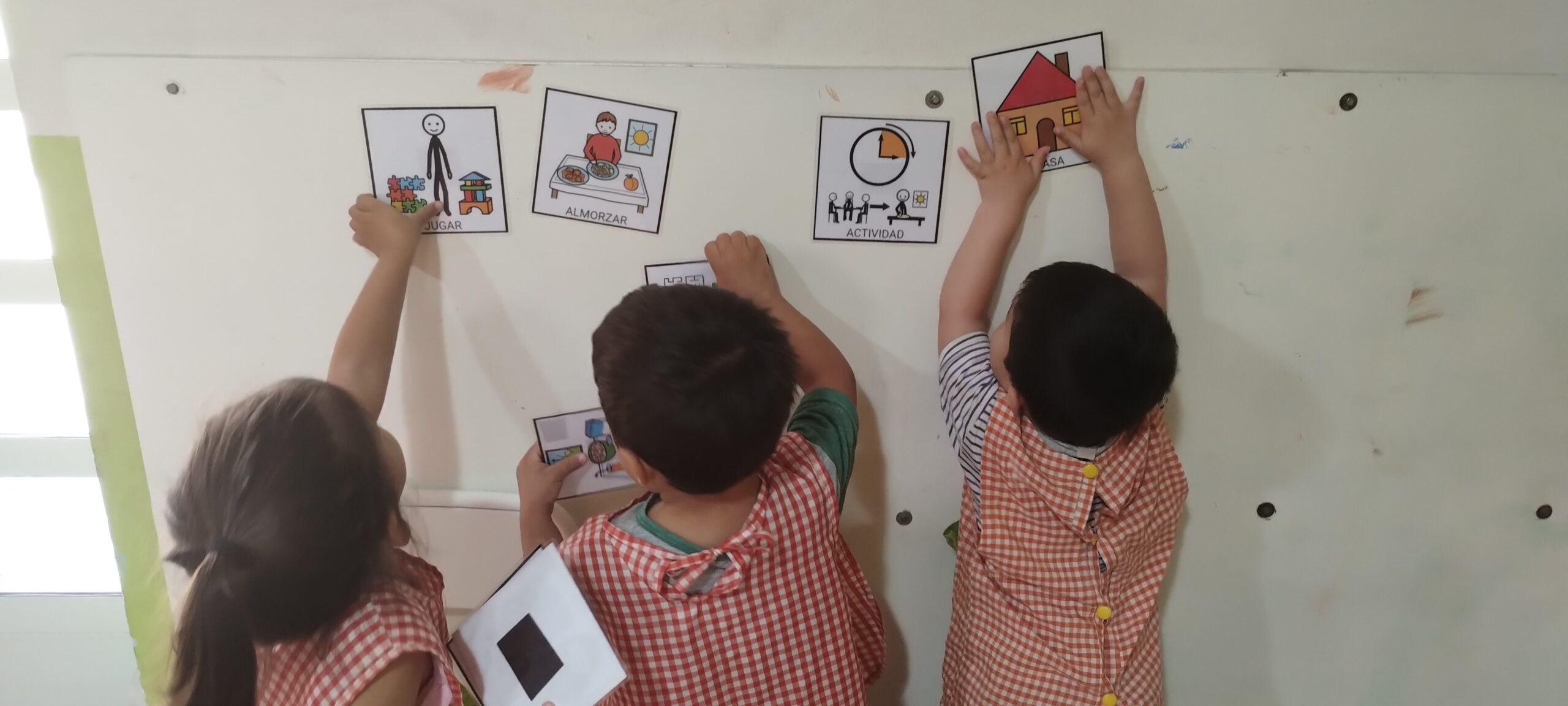 La imagen muestra a tres niños pequeños, vistos desde atrás, vestidos con delantales de cuadros rojos y blancos. Están frente a una pared blanca,organizando su rutina a través de pictogramas.