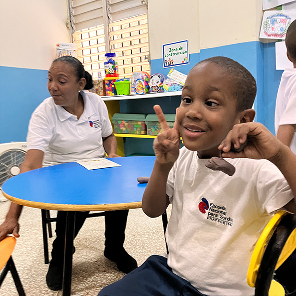 Un niño sonríe y hace el signo de la paz sentado en la mesa de clase. Una maestra está sentada cerca, mirando hacia otro lado. El aula tiene juguetes de colores y adornos en las estanterías del fondo.