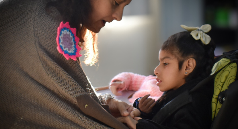 Una mujer se inclina hacia una niña en silla de ruedas y le coge suavemente la mano. La niña lleva un lazo blanco en el pelo y parece concentrada, mientras que la mujer parece cariñosa y atenta. Una luz suave ilumina sus rostros.