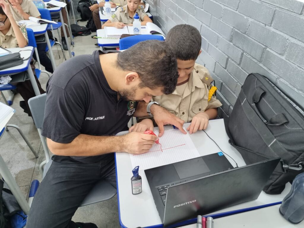 Um professor ajuda Pedro, um jovem, aluno a fazer o trabalho escolar em uma mesa, desenhando em um papel pautado. Ambos usam uniformes. Um laptop, um frasco de tinta e um estojo de lápis estão sobre a mesa. Outros alunos trabalham ao fundo.