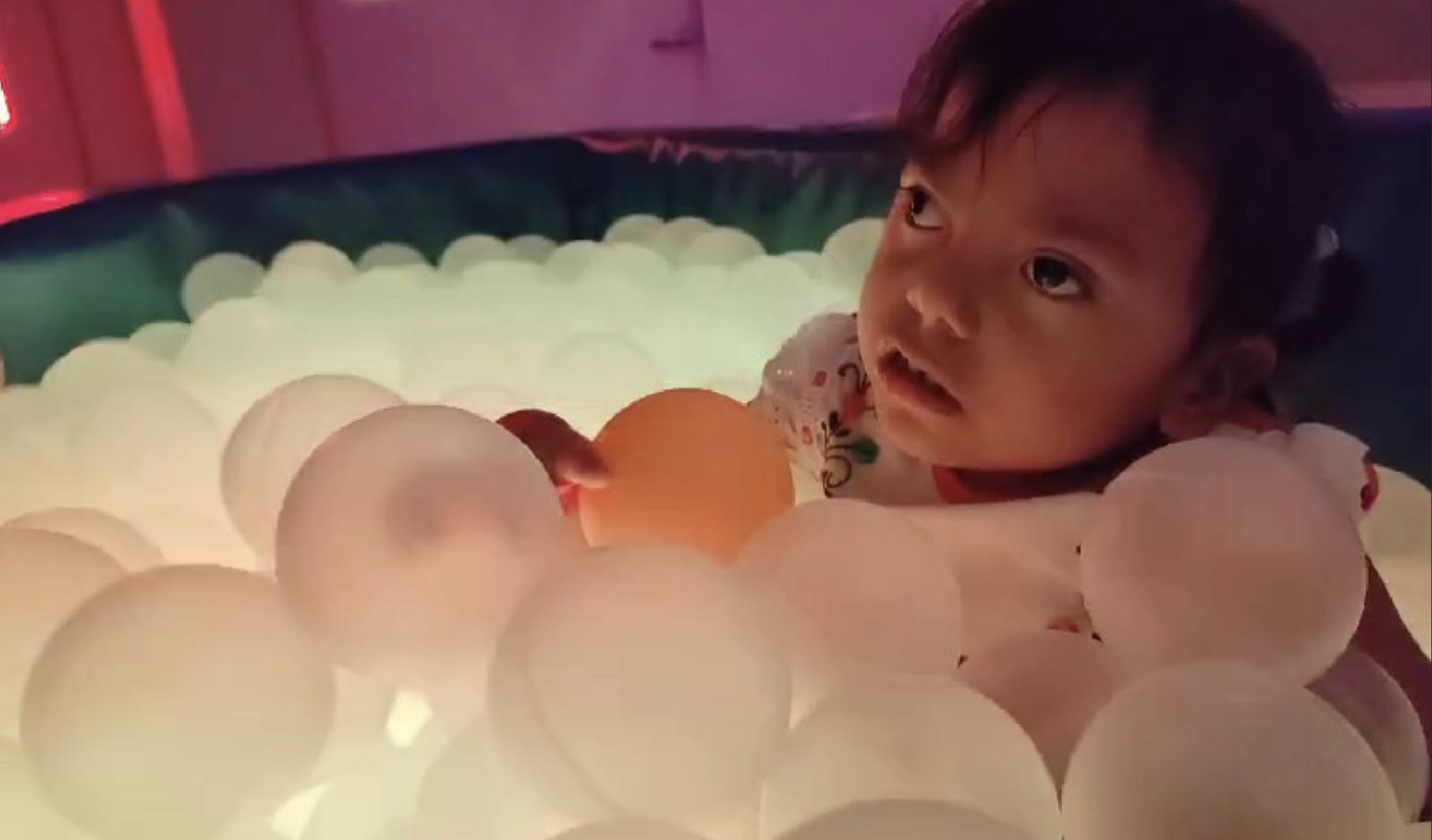 Una niña pequeña está inmerso en una piscina de bolas, mirando hacia arriba con curiosidad. Las bolas de plástico que lo rodean son en su mayoría blancas, con algunas de colores, y la iluminación da a la escena un brillo cálido y suave.