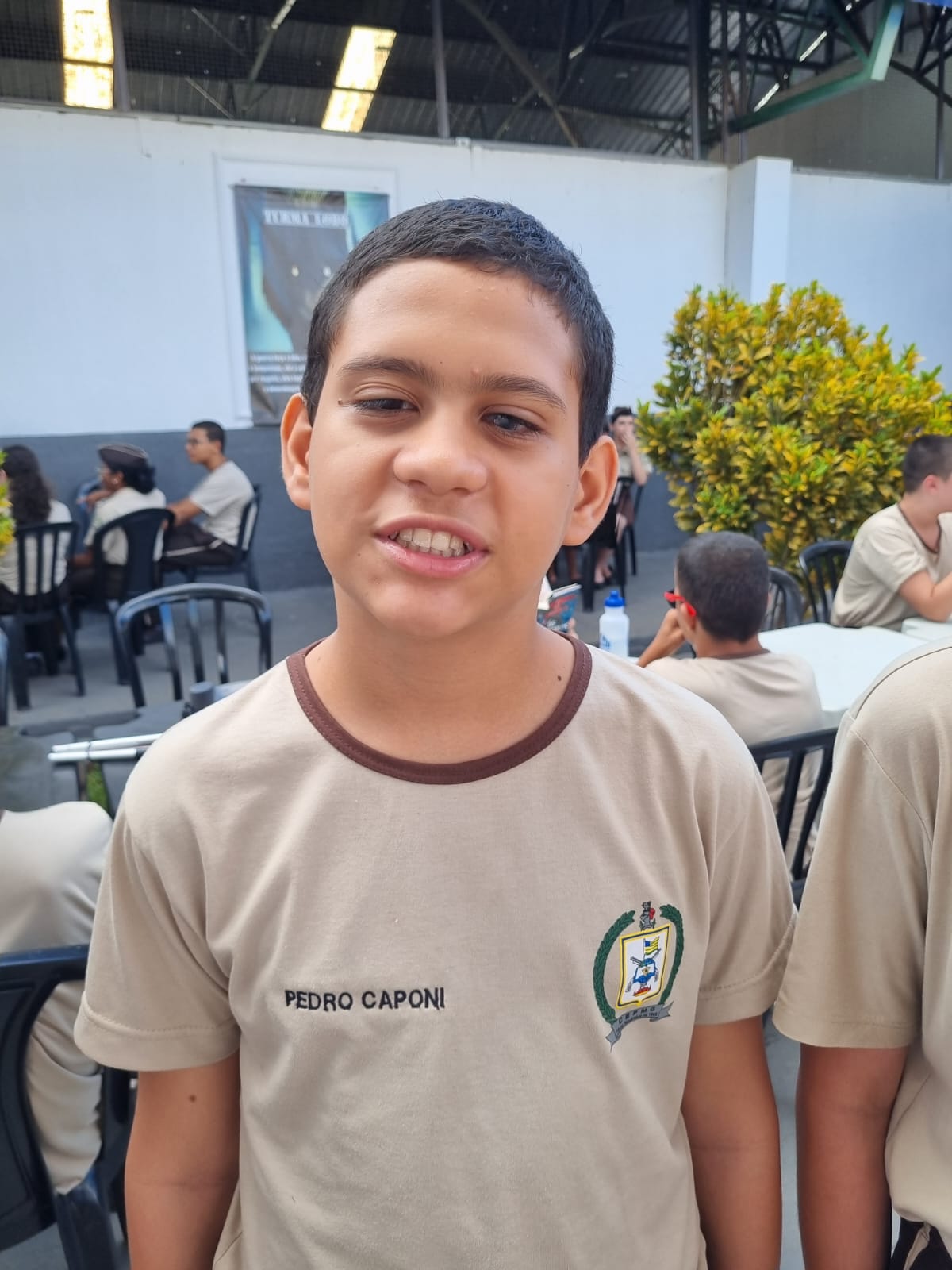 Un joven sonriente, vestido con una chaqueta negra y beige con la inscripción "98" en la parte delantera, está sentado en un banco de madera contra una pared verde brillante.