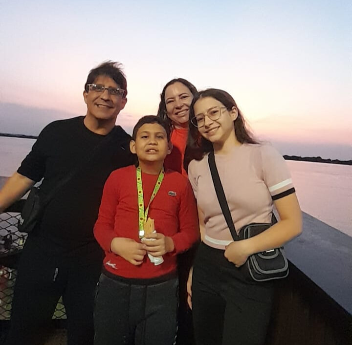 Pedro, os seus pais e a sua irmã estão juntos do lado de fora, sorrindo para a câmara. Atrás deles há um corpo de água e o sol está a se pôr, tornando o céu e a água cor-de-rosa.