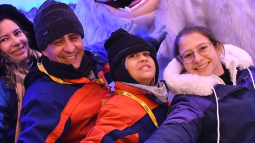 Pedro, os seus pais e a sua irmã, todos vestidos com casacos e chapéus de inverno, sorriem e posam juntos em frente a uma grande estátua de uma criatura de neve, sugerindo um passeio divertido num ambiente nevado ou temático.